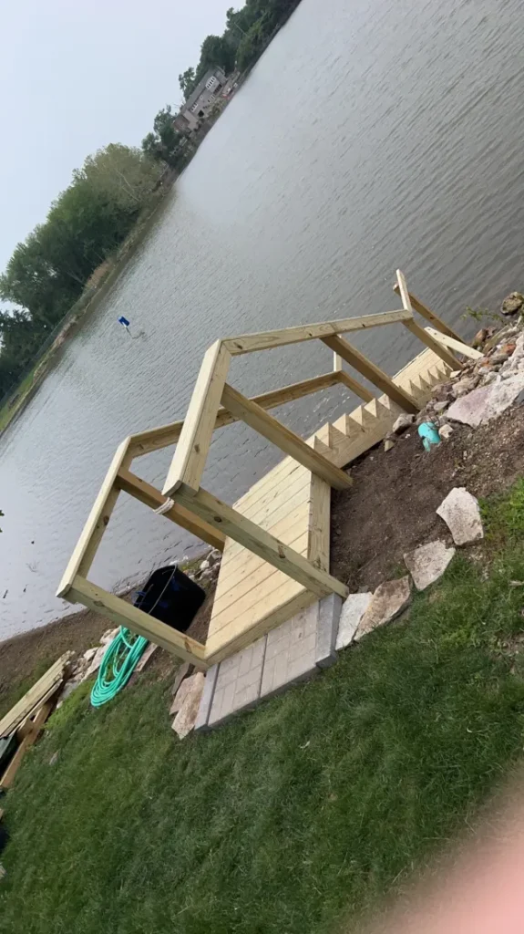 Unfinished wooden dock structure extending into a body of water from a grassy bank.