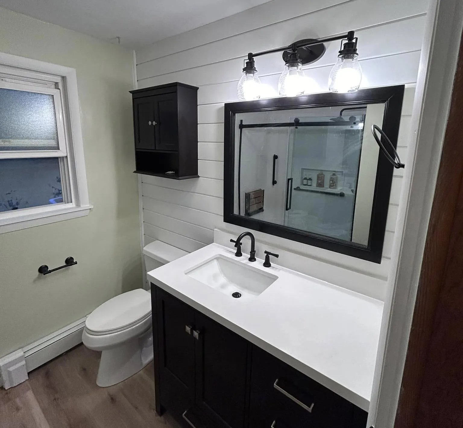 A newly renovated bathroom with light green walls, a black vanity with a white countertop and sink, a black framed mirror, and a black three-light vanity fixture.