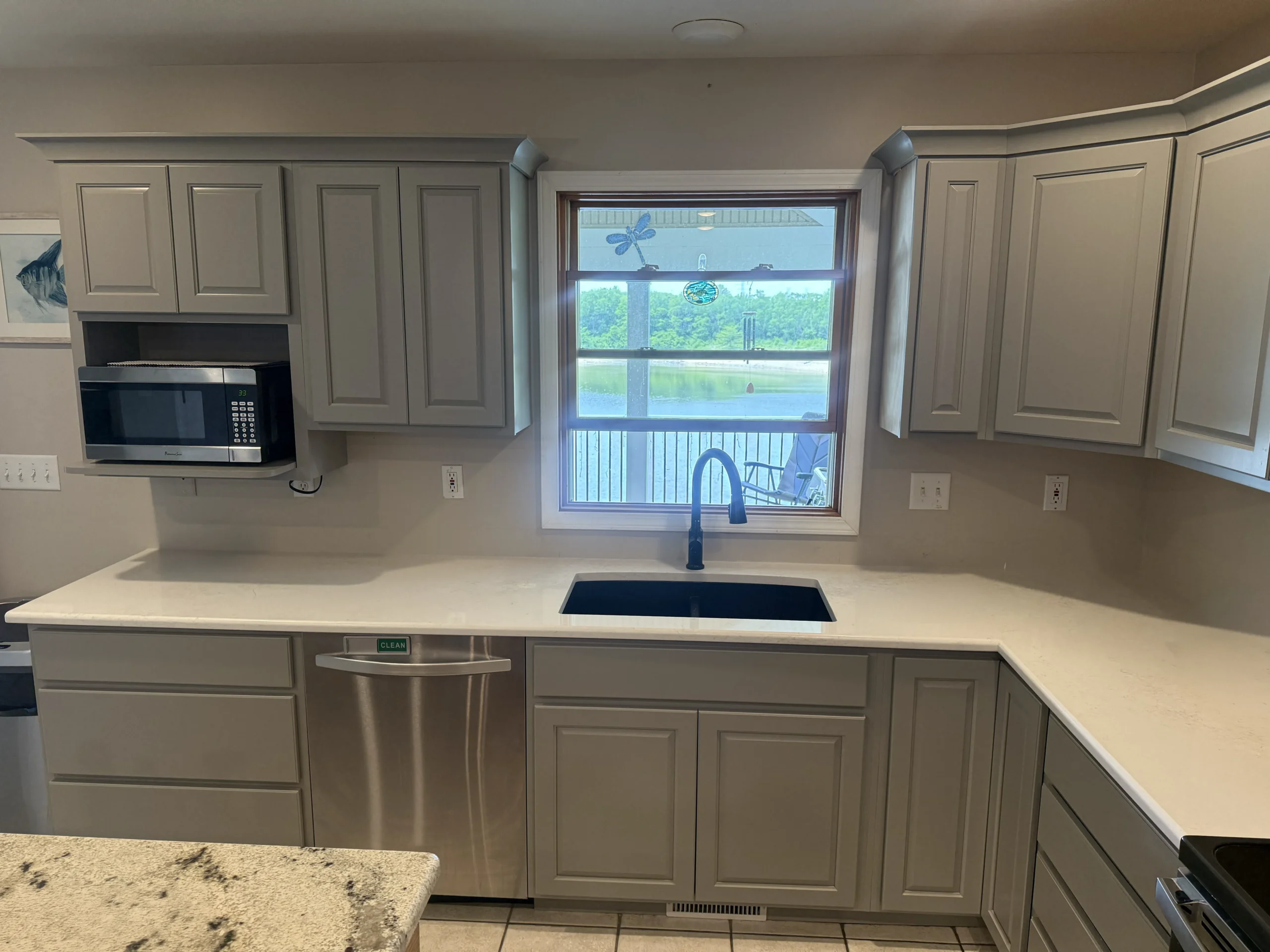 A recently renovated kitchen with light grey shaker style cabinets, white countertops, and a black sink with a black faucet positioned under a window offering a view of a body of water and trees, Creve Coeur, IL Top Construction.