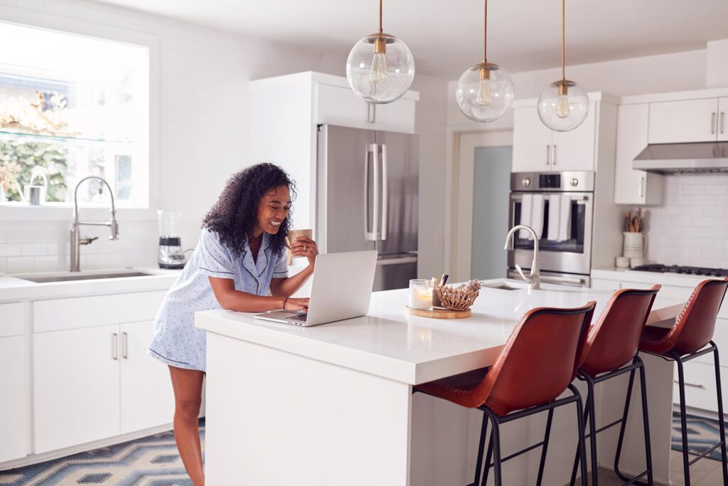 Woman in pajamas smiling and working on laptop in a modern kitchen with white cabinets and a large island
