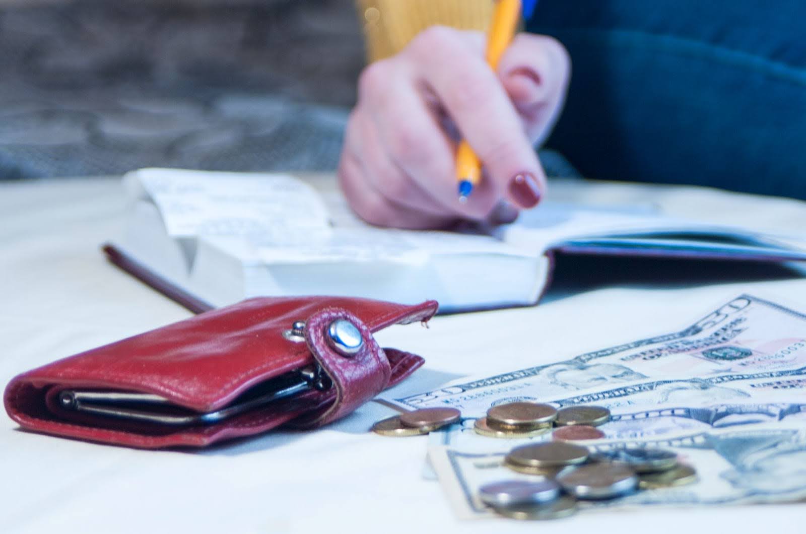 Woman writing a home remodel budget in a notebook with a red wallet and cash beside it on the table.