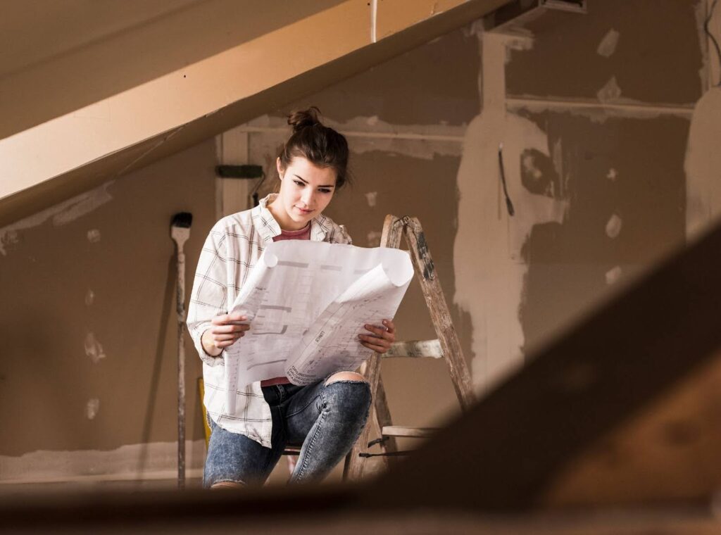A young woman is sitting on a ladder looking over a blue print.