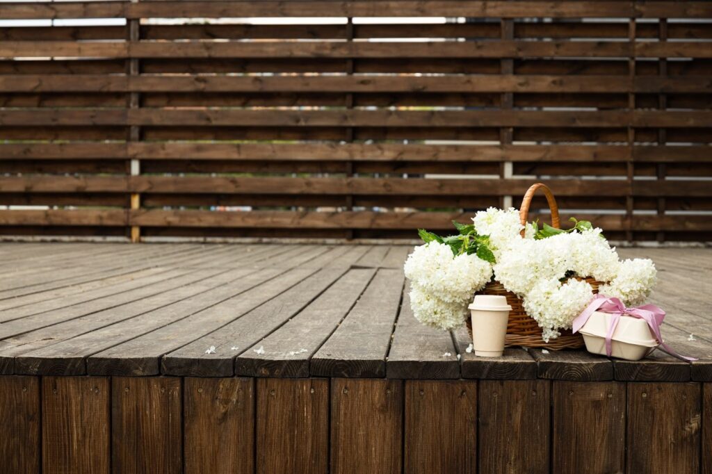 Flowers, a cup, and a to-go box of food sit on a wooden deck.