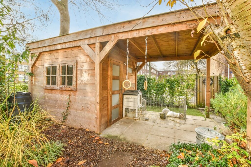 Country-style backyard junior accessory dwelling unit with a covered veranda and a dog resting safely inside a cage.