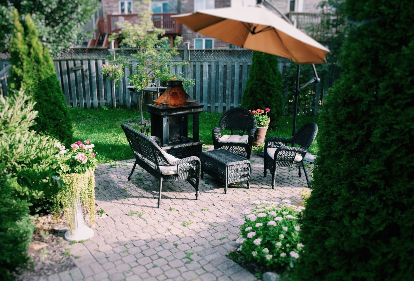 Small backyard patio featuring a compact metal fireplace and black wicker seating set, surrounded by garden greenery.