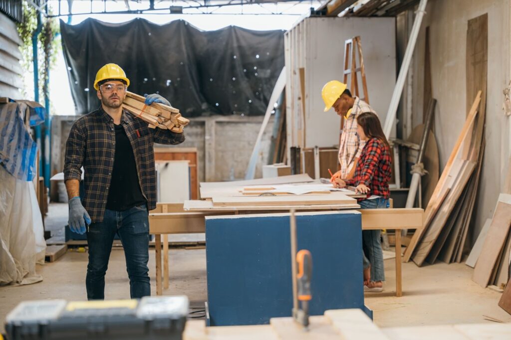 Carpenter carrying wood planks on his shoulder, wearing a safety helmet and gloves, while the architect and engineer review renovation plans on-site.