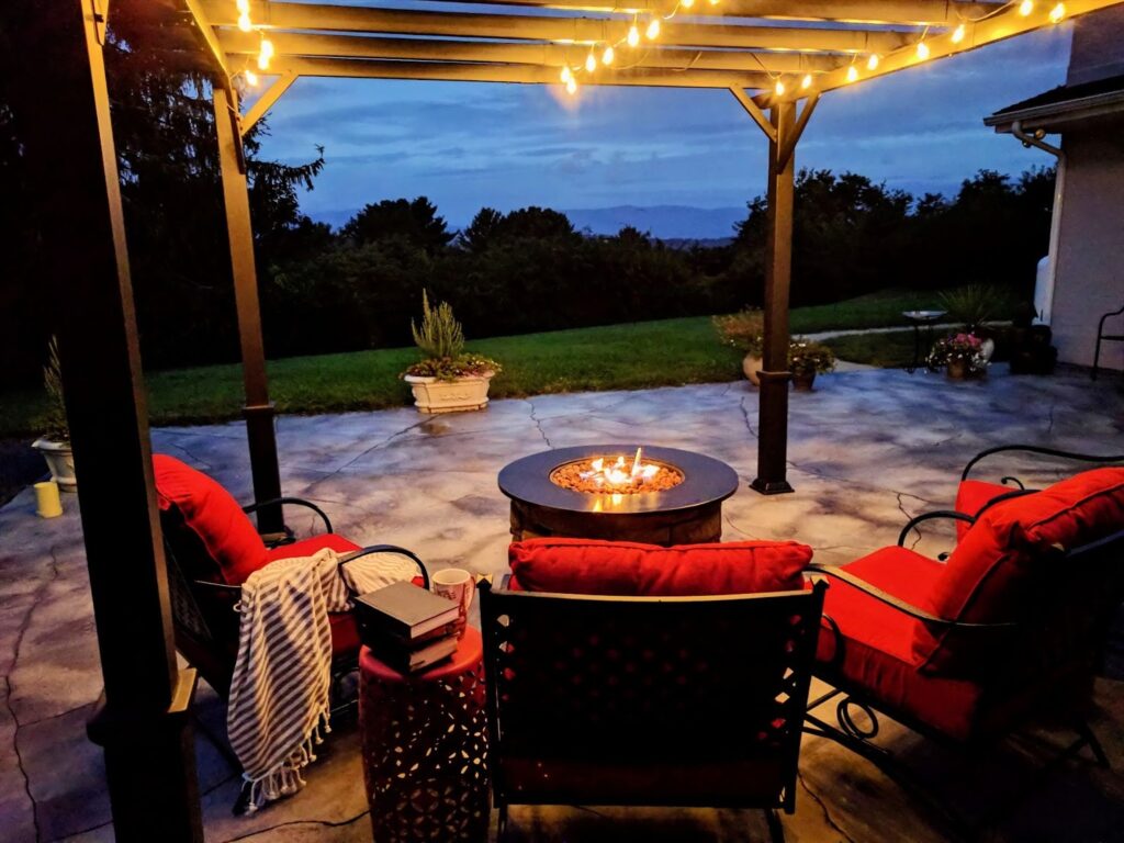 Outdoor living space at dusk featuring a gas fire pit, red cushioned seating, and warm string lights hanging from a slatted patio cover..