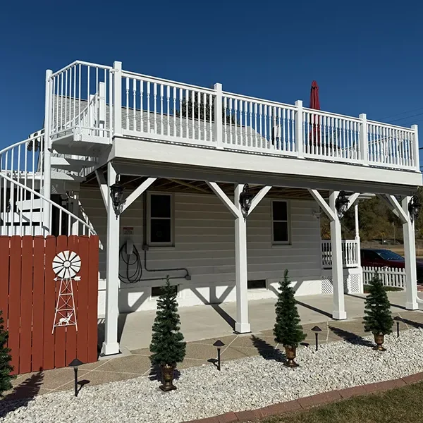 White elevated house with a spacious rooftop deck and ground-level patio with small evergreen trees.