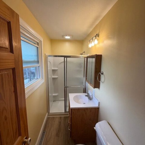 A small, narrow bathroom with yellow walls, a wooden vanity with a white sink, a toilet, and a glass shower stall.