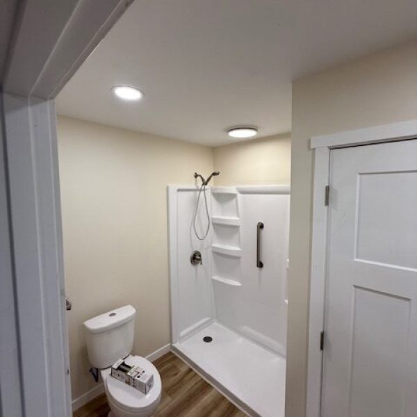 A modern bathroom with a white shower stall, a toilet, recessed lighting, and a white door with a window pane design.