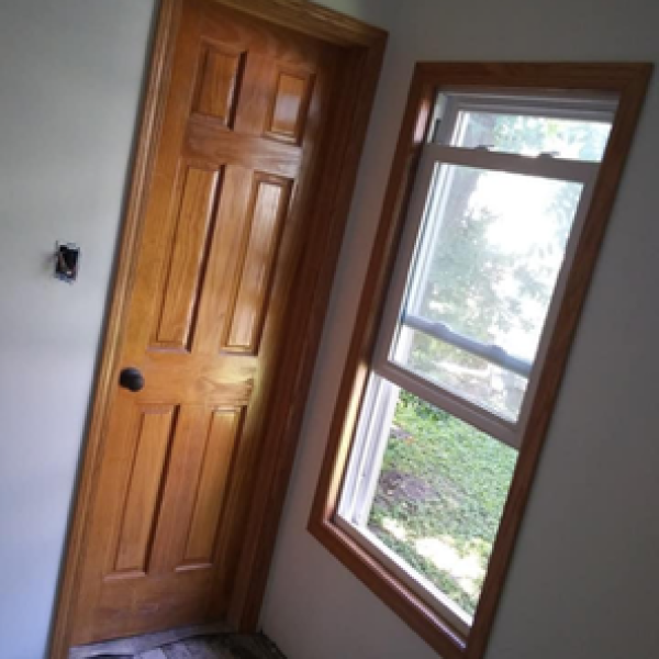 Corner of a room showing a newly installed wooden six-panel door and a window with a wooden frame.
