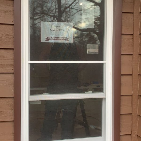 A close-up of a new Simonton double-hung window with white frames and brown trim on a house with tan siding.