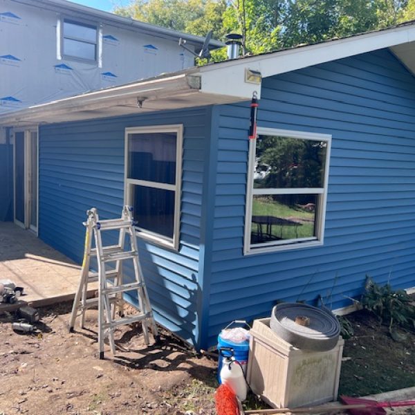 An exterior view of a house addition under construction, featuring new blue vinyl siding, newly installed windows, and a ladder on a dirt patio.